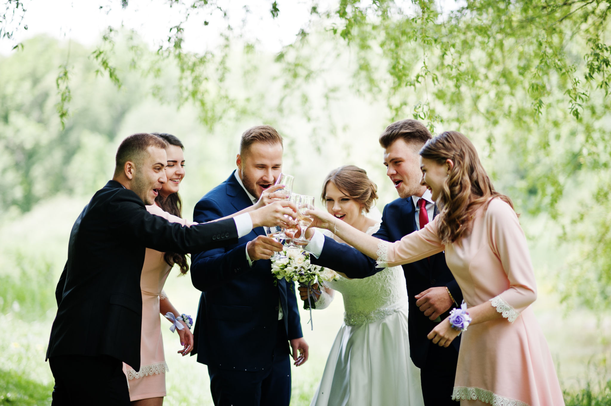 Wedding couple, bridesmaids and groomsmen drinking champagne on a festive wedding day in the park.